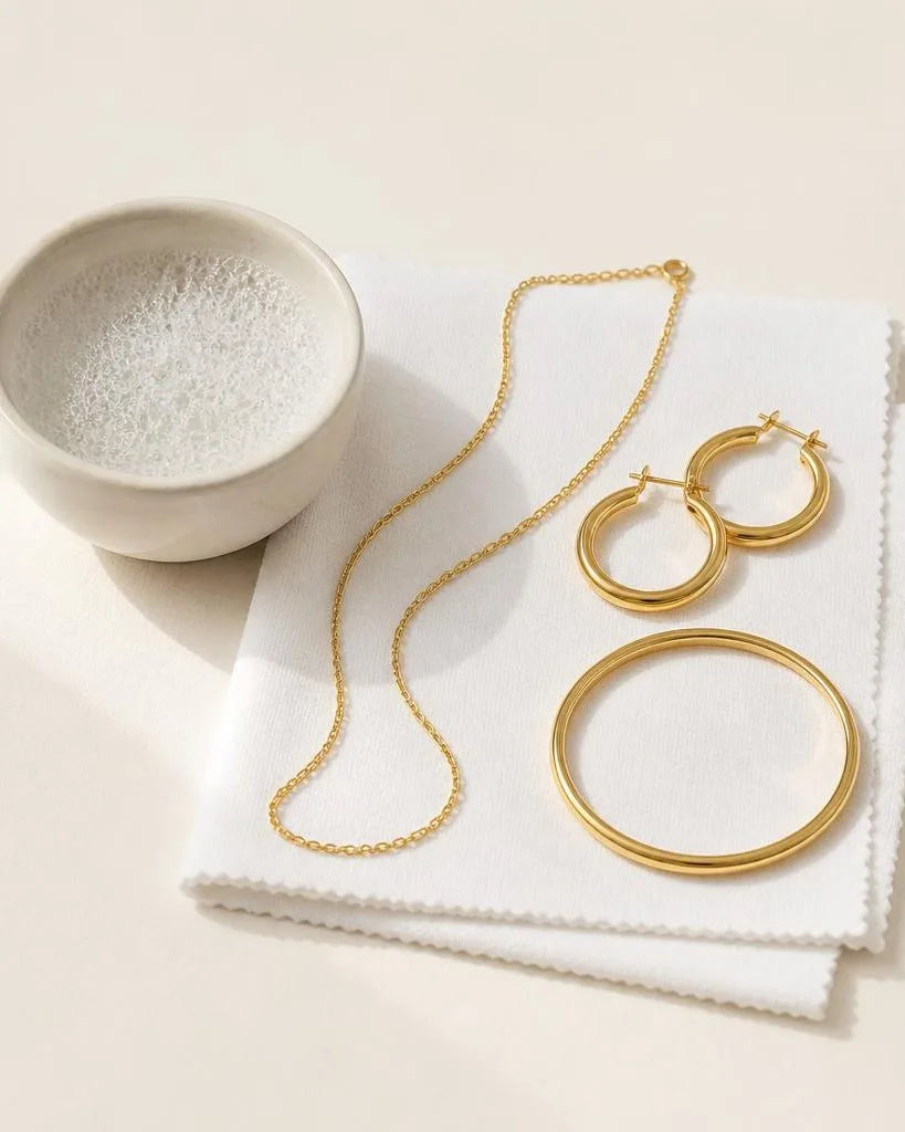 Bijoux plaqués or posés sur un chiffon microfibre à côté d'un bol d'eau savonneuse, prêts pour un nettoyage doux