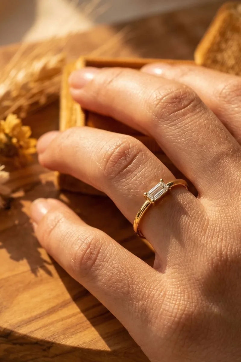 18K yellow gold baguette cut diamond solitaire ring worn on the finger against a natural background, by Mayuri